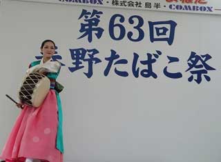 「第63回 秦野たばこ祭」と大書された白い看板の前で、ピンクと水色のチマチョゴリを着用し、韓国の伝統的な太鼓であるチャングを抱えて、笑顔でポーズをとる女性の演者の写真