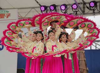 屋外のステージで、チマチョゴリを着用した数名の女性たちが、赤とピンクの華やかな扇子を頭上に掲げて円形に広げる韓国の伝統舞踊を披露している様子の写真