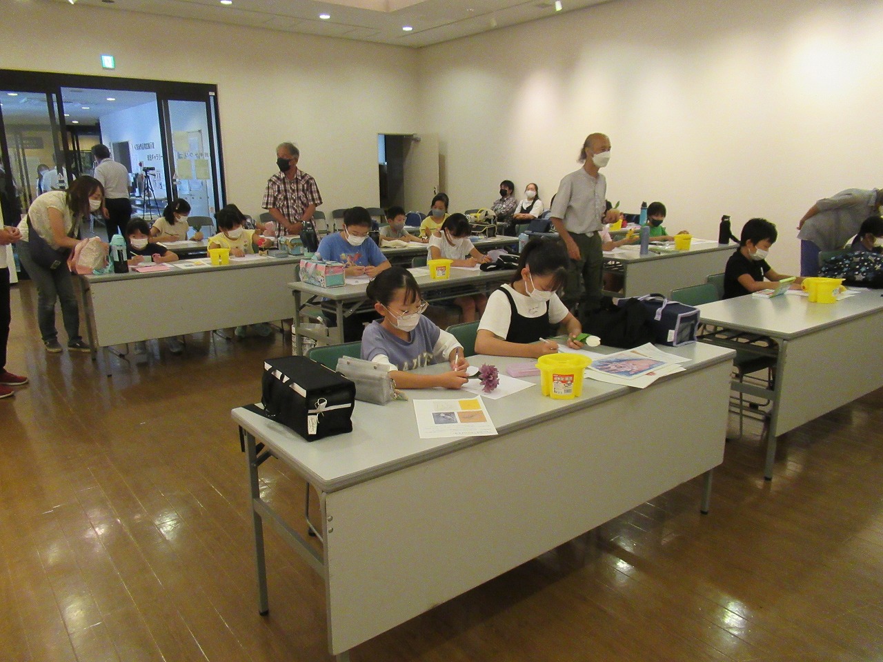 大勢の子供たちが黄色のバケツや道具が置かれた机に向かって座り、マスクを着用した講師たちが指導する中、絵を描いたり色を塗ったりしている様子の写真