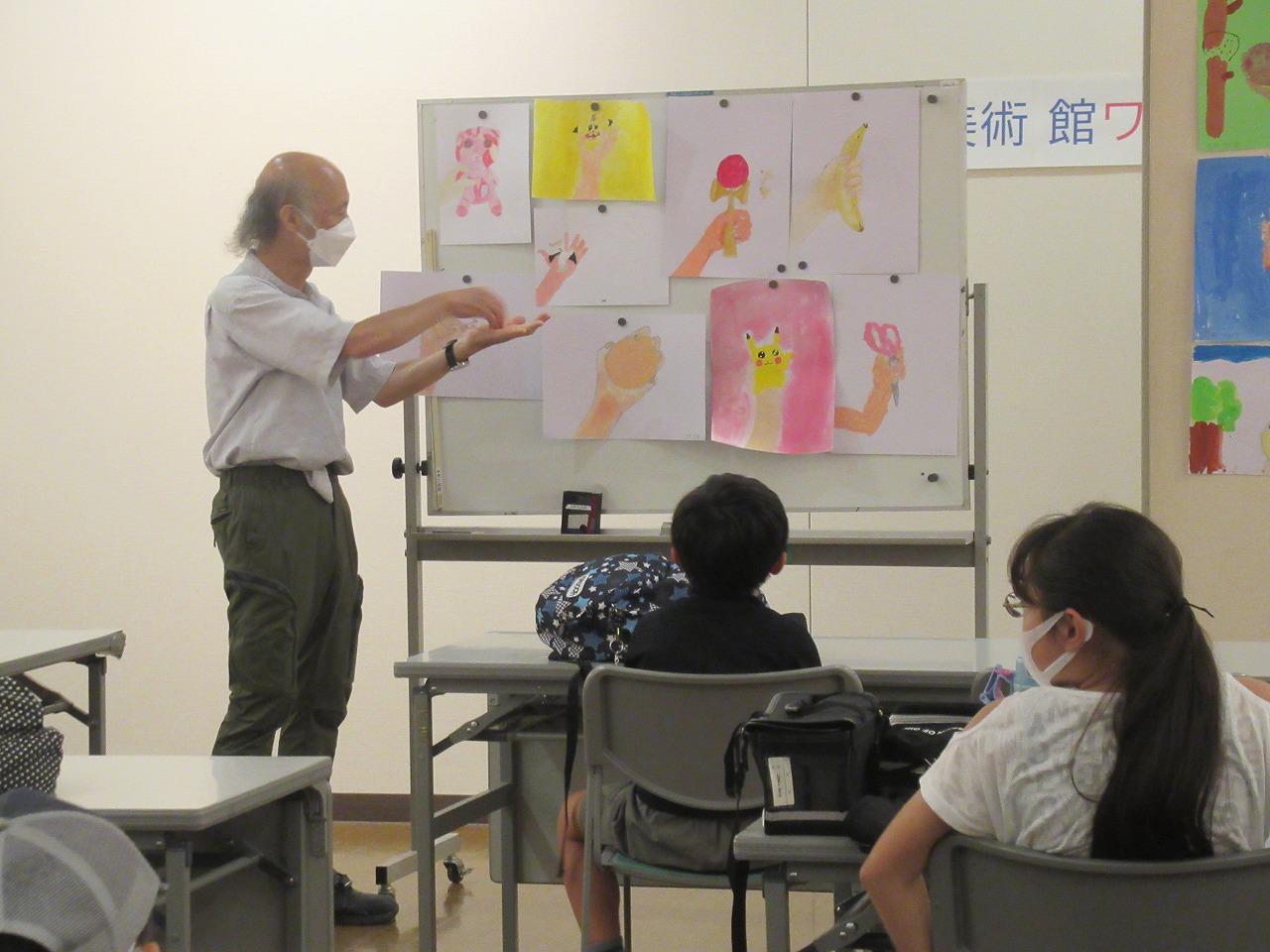 白いホワイトボードに子供たちが描いたぬいぐるみやバナナなどを持った手の絵が複数枚展示されており、男性講師が身振り手振りで、マスクを着けた子どもたちに熱心に説明している写真