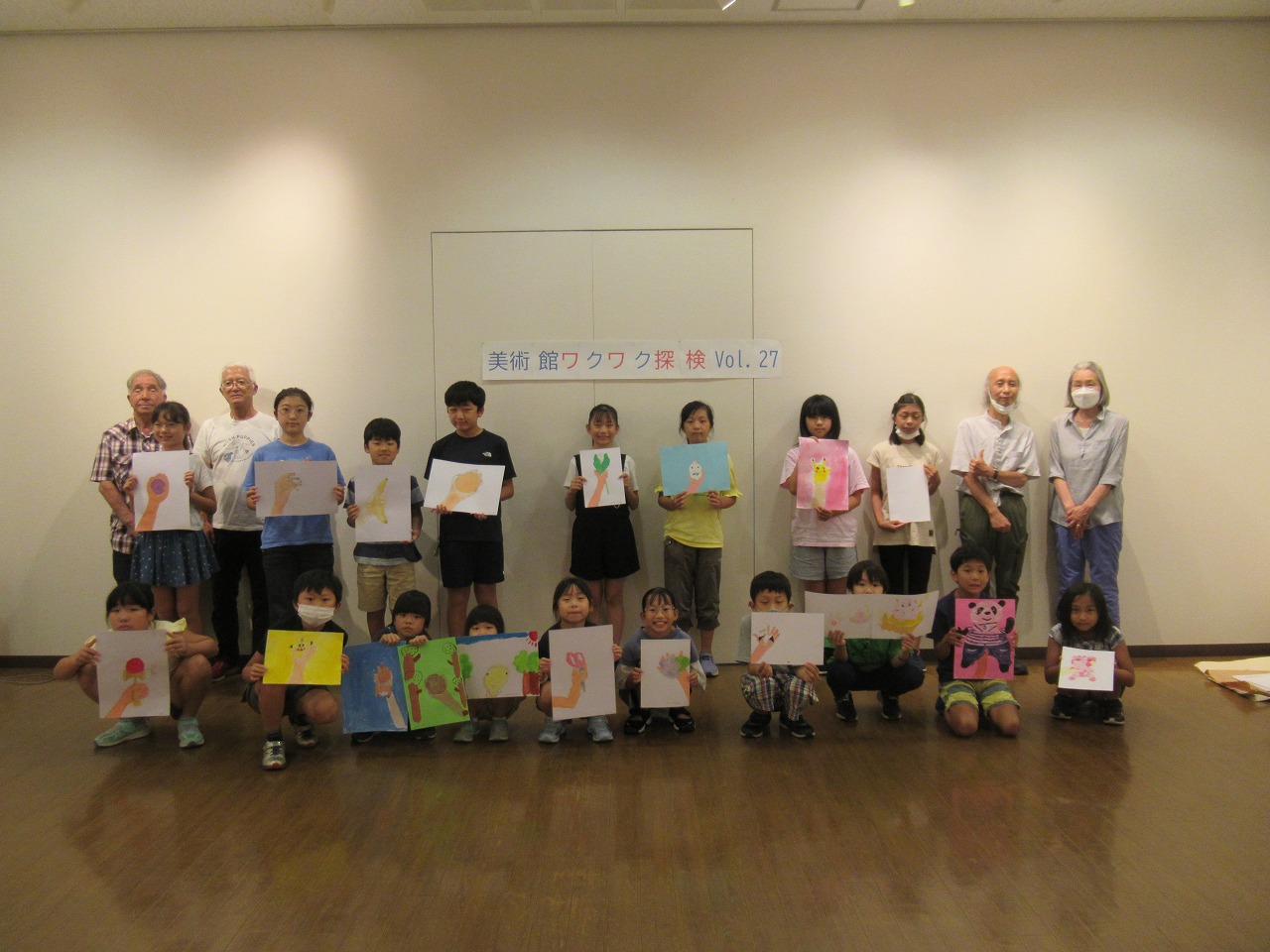 「美術館ワクワク探検 Vol.27」と書かれた壁の前で、子供たちが描いたそれぞれの絵を手に持ち、4人の講師と共に記念撮影をしている集合写真