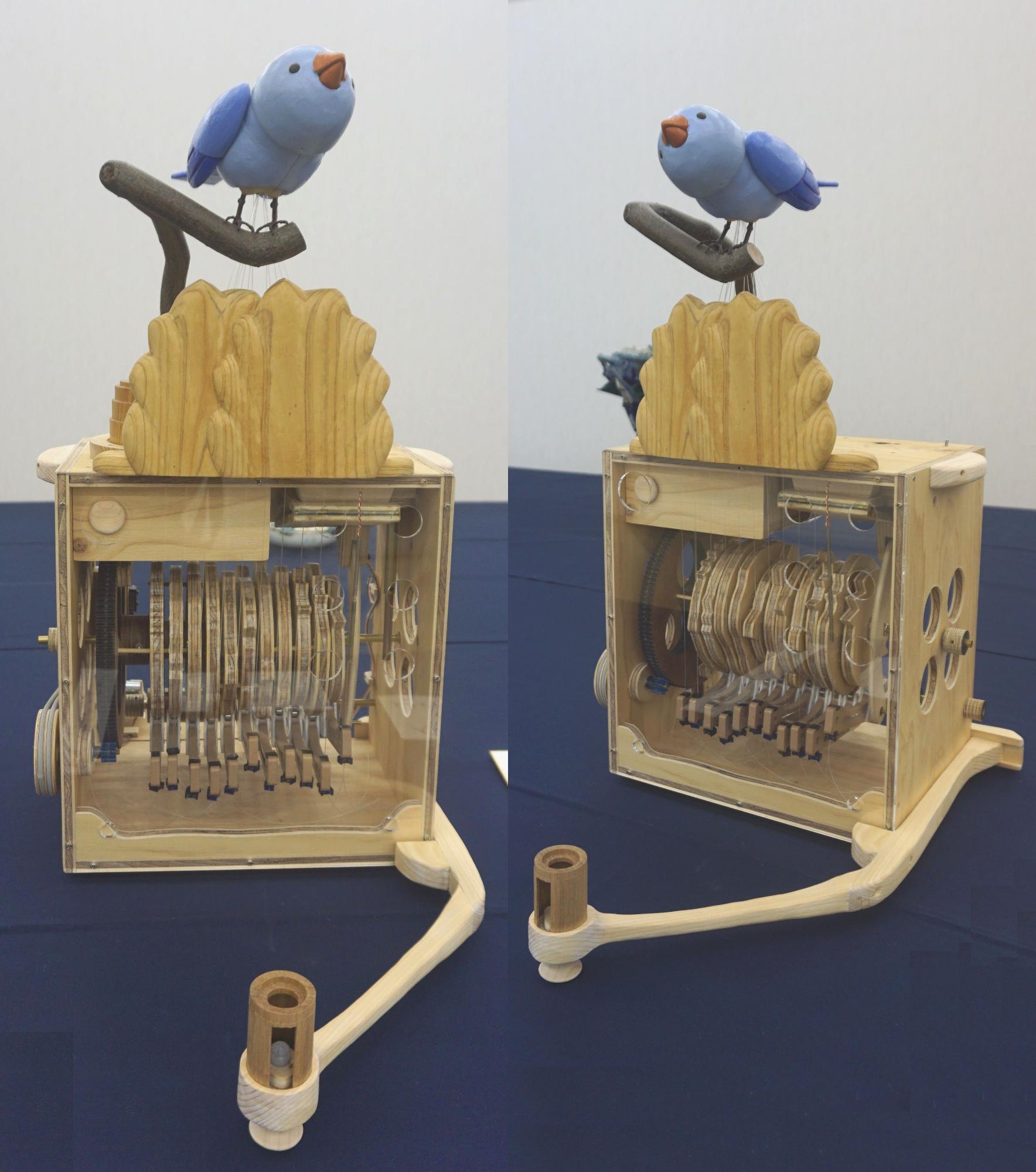 正面と右斜めから撮影された木製のからくりおもちゃで、青い鳥がとまった台座の下にある正方形の透明の箱の中には複雑な歯車と鍵盤のような機構が見える、美術の部で市展大賞を受賞した作品の写真