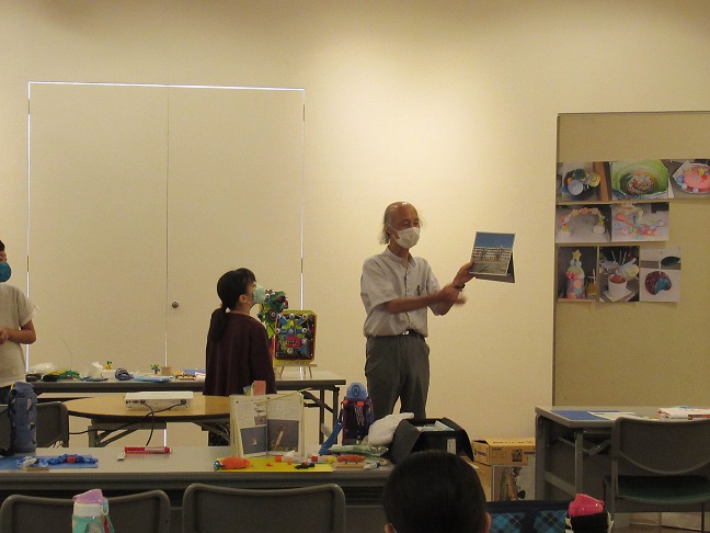 沢山の絵が貼られた掲示ボードの傍に立つマスクをつけた白いシャツ姿の男性講師が、子どもたちに向かって学校の校舎の写真のようなものを見せながら作品の説明をしている写真