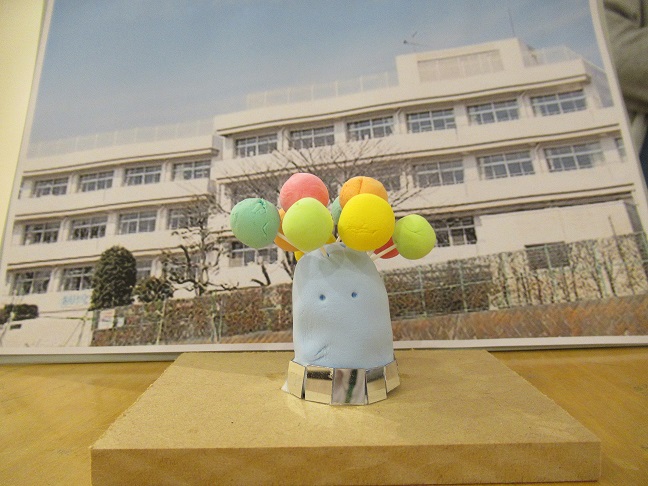 目がついた淡い水色の紙粘土に色とりどりの円形の球体が刺さっており、底の部分に銀紙で装飾が施されているオブジェが、学校の写真を背景にして置かれている子供の作品の写真