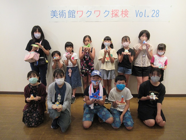 「美術館ワクワク探検 Vol.28」と書かれた壁の前で、参加した子供たちがそれぞれ制作した粘土細工を手に持ち、マスクを着用して2列に並んでいる集合写真