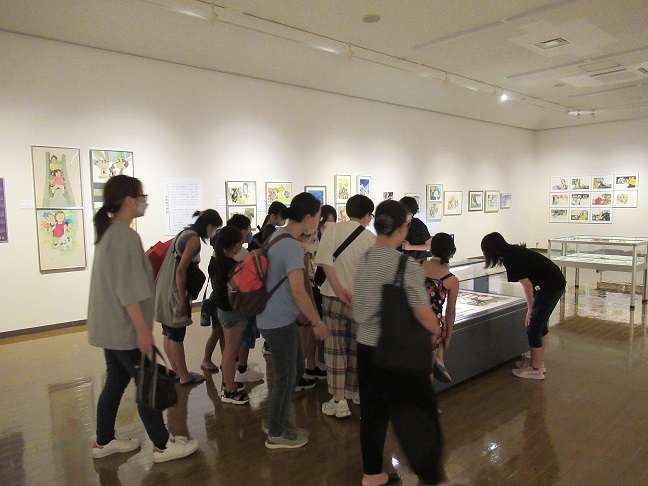 白い壁に子供向けと思われるイラストや絵画が複数展示されている美術館内で、中央のガラスケースの周りに集まった大勢の親子連れが展示物を鑑賞している様子の写真