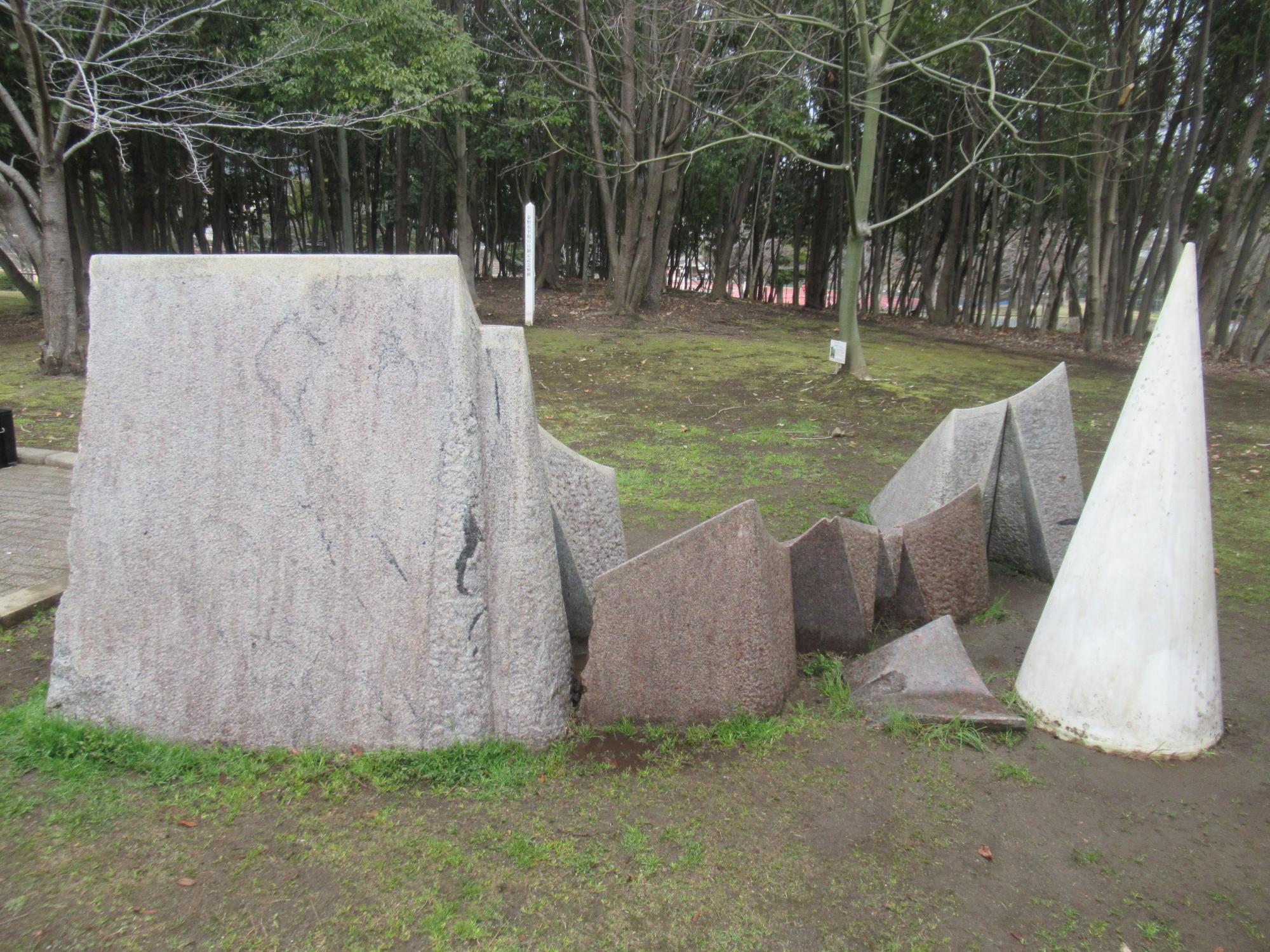 背景には細い木々が立ち並ぶ広場に、いくつかの大きな石の彫刻が並んでおり、左側には四角形に近い灰色の石があり、その隣には多角形の石が複数配置され、右端には白い円錐形の石が立っている彫刻群の写真