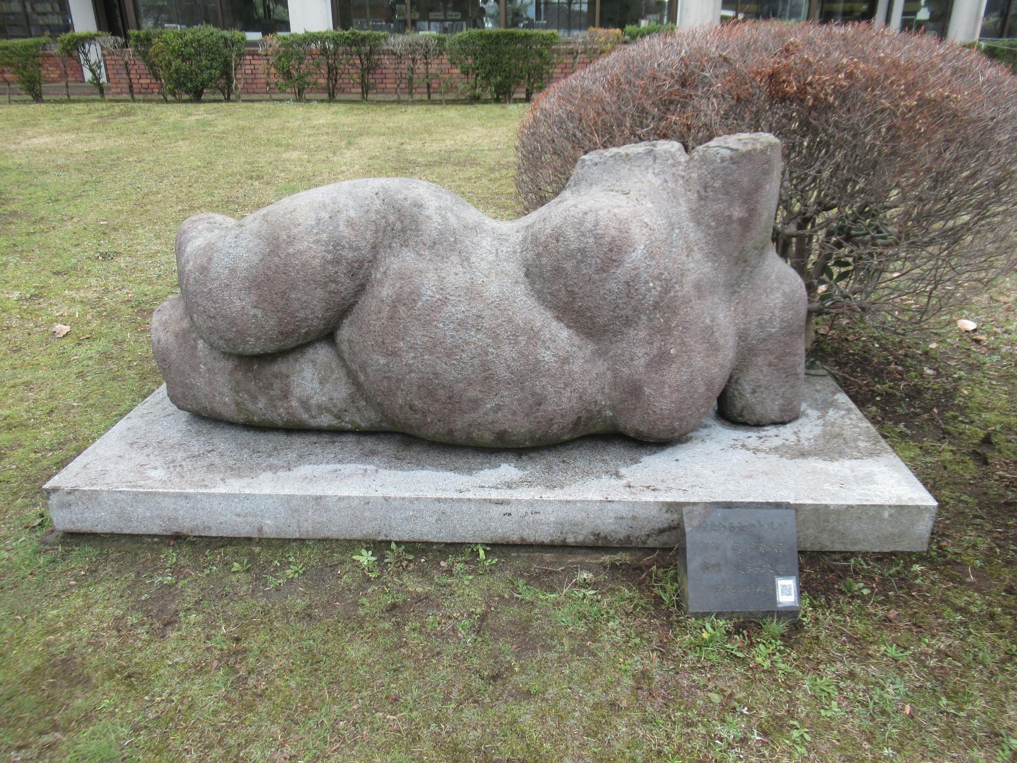 レンガ調の外壁の建物の前にある芝生広場に、四角い台座の上に、頭部と両腕、両脚部を欠いた横たわる肉付きの良い女性のトルソを思わせる、抽象的な石の彫刻が置かれた野外アート作品の写真