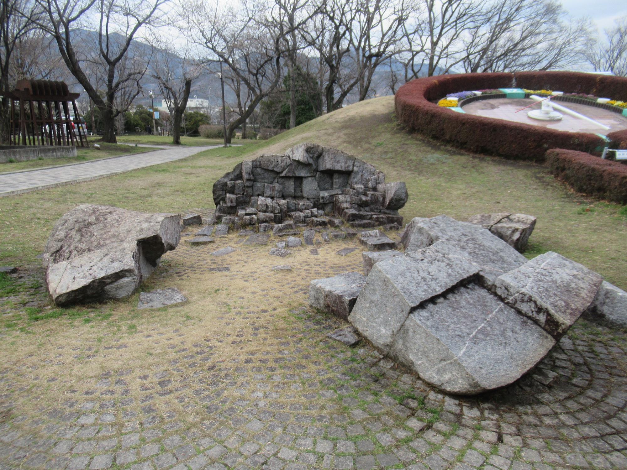 木々に囲まれた小高い広場に設置された花時計のある手前の平坦な石畳の上に、砕けた不揃いな形をした大きな岩が複数配置され、中央には積み重ねられた石ころの構造物が見える彫刻の写真