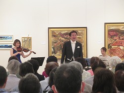 絵画が飾られた白い壁の会場で、多くの聴衆が座って鑑賞しているなか、右側で女性がピアノを弾き、左側で別の女性がバイオリンの演奏に合わせて、中央に立つタキシードを着た男性歌手が歌っているコンサートの様子の写真