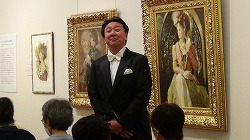 豪華な額縁に入った肖像画が飾られている会場で、タキシードを着て蝶ネクタイをした男性が、美術館の展示室で来場者を前に立っている写真