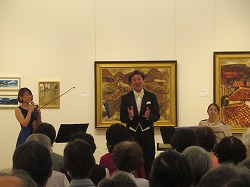 絵画が展示されたホールで、左に立ってヴァイオリンを持つ女性と右のピアノの前にを座っている女性の間で、タキシードの男性が中央で歌っているコンサートの様子を捉えた写真