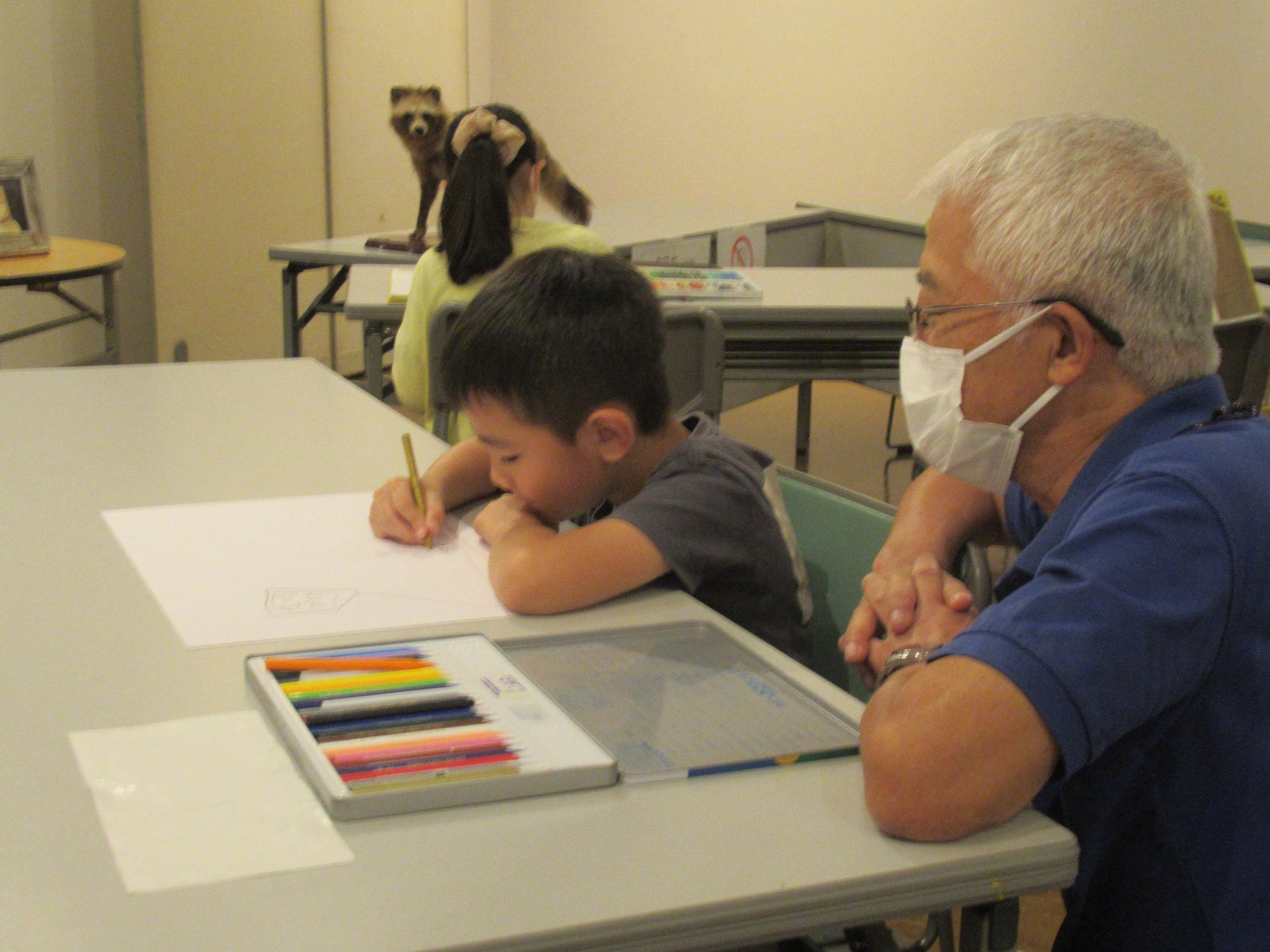 白いマスクを着けた年配の男性が見守っているなか、色鉛筆のセットが置かれた机に向かう男の子たスケッチを行っているワークショップの様子の写真
