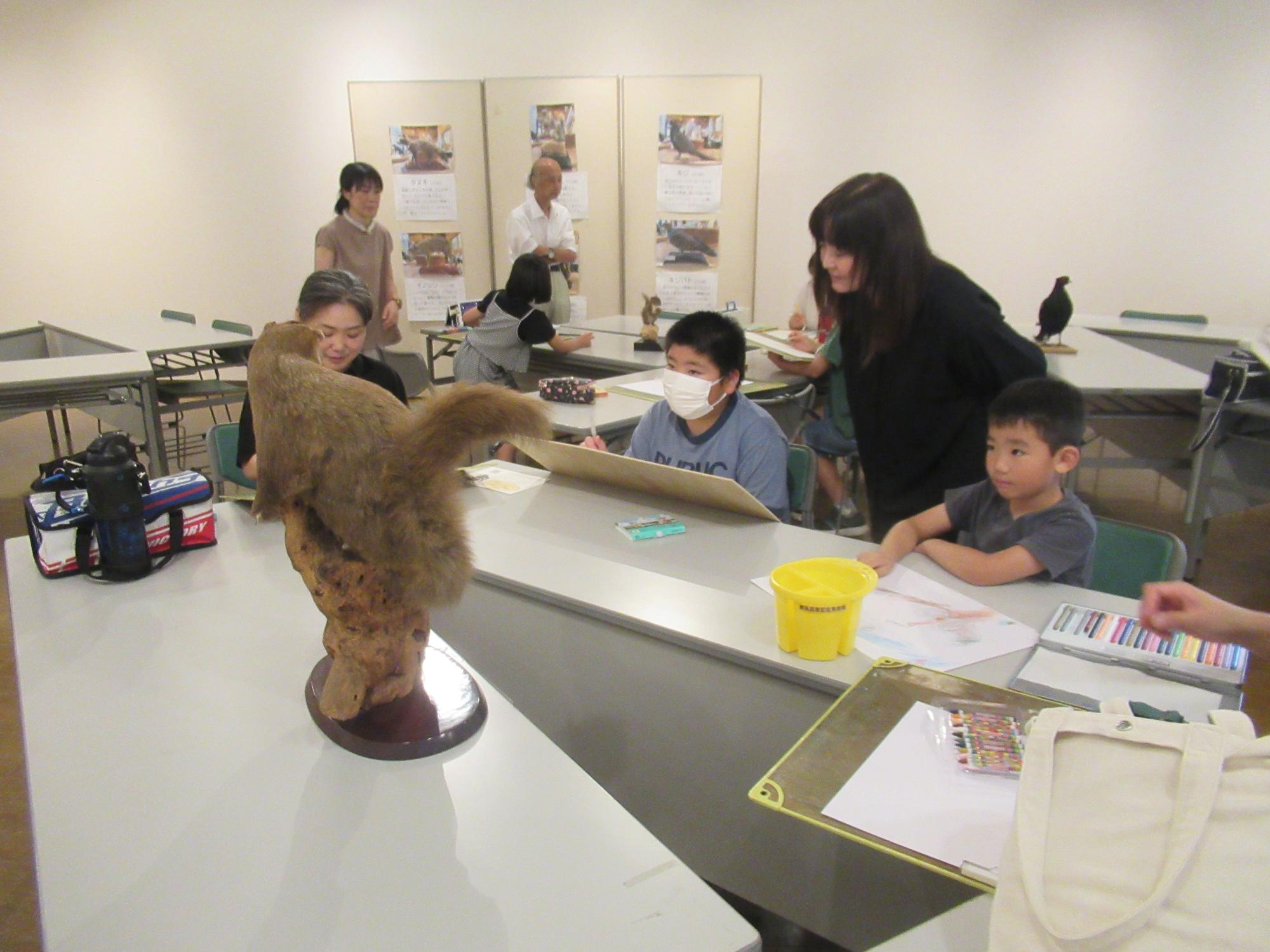美術館の創作室で、女性講師が見守る中、3人の子供たちが机の上にある動物の剥製を観察しながら絵を描いている様子の写真