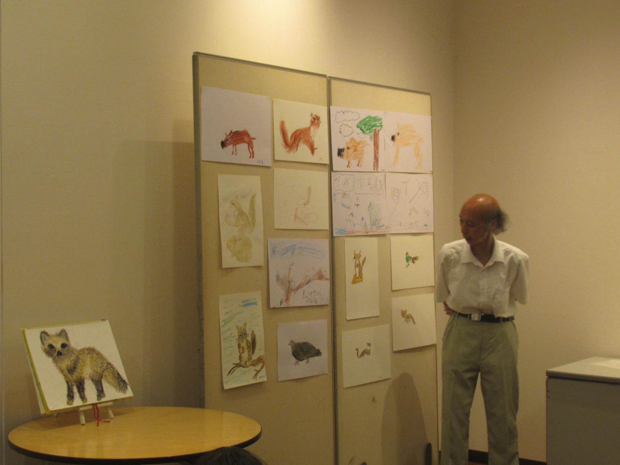 部屋の壁側に設置された2枚の掲示ボードに、子供たちが描いた動物の絵画が複数枚展示されており、傍に立つ白いシャツを着た男性講師が話をしている様子の写真