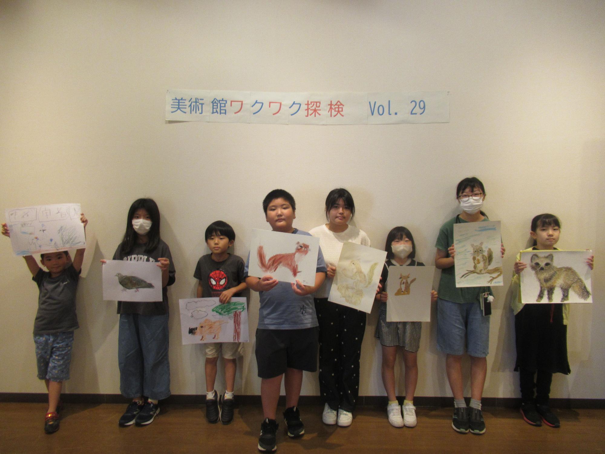「美術館ワクワク探検 Vol.29」と書かれた壁の前で、8人の子供たちがそれぞれ制作した動物の絵を手に持ち、皆で記念撮影をしている集合写真