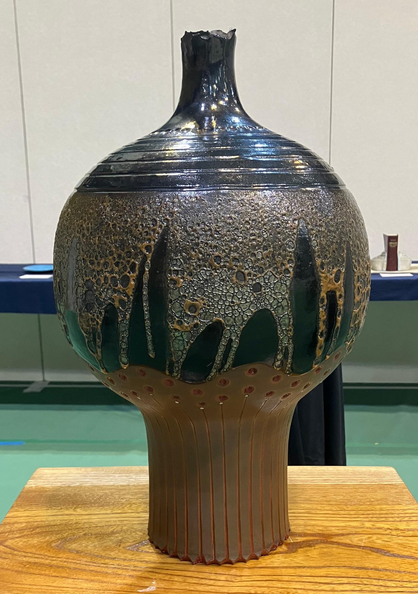 どっしりとした胴と細く伸びた首を持つ陶器の大きな花瓶が木製の台座の上に置かれ、胴の上半分には光沢のある黒と茶色の垂れた釉薬がかけられている、美術の部で市展大賞を受賞した作品の写真
