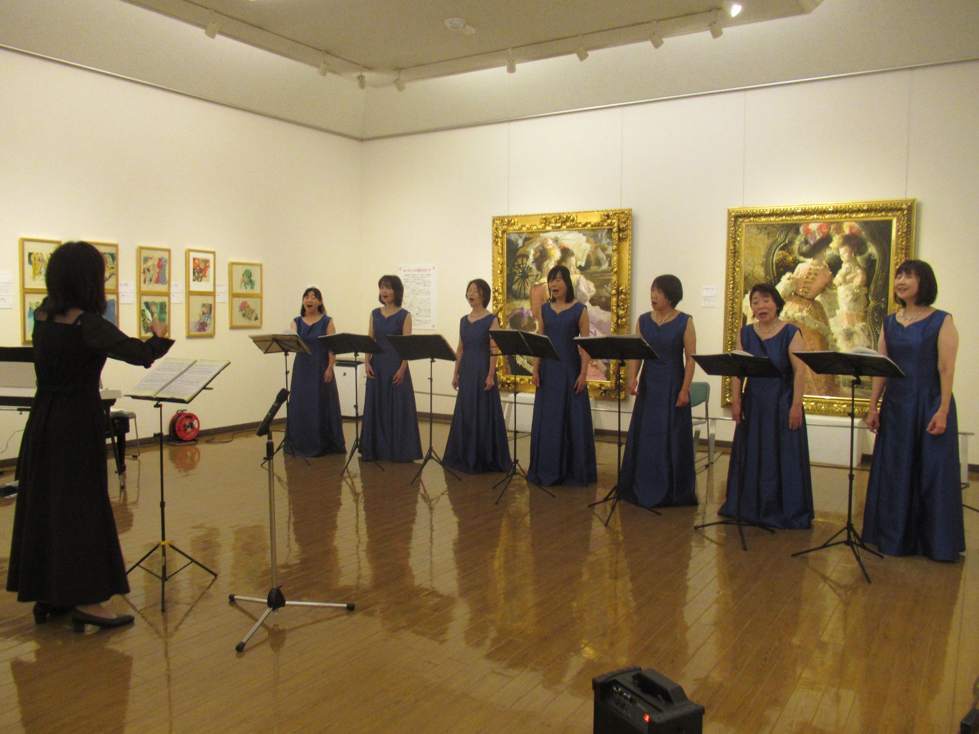 背景には豪華な額縁の絵画がいくつも飾られている美術館の展示室で、譜面台の前に立つ黒いドレスの指揮者に合わせて濃紺のロングドレスを着た7人の女性が歌を披露している様子を右斜めから撮影した写真