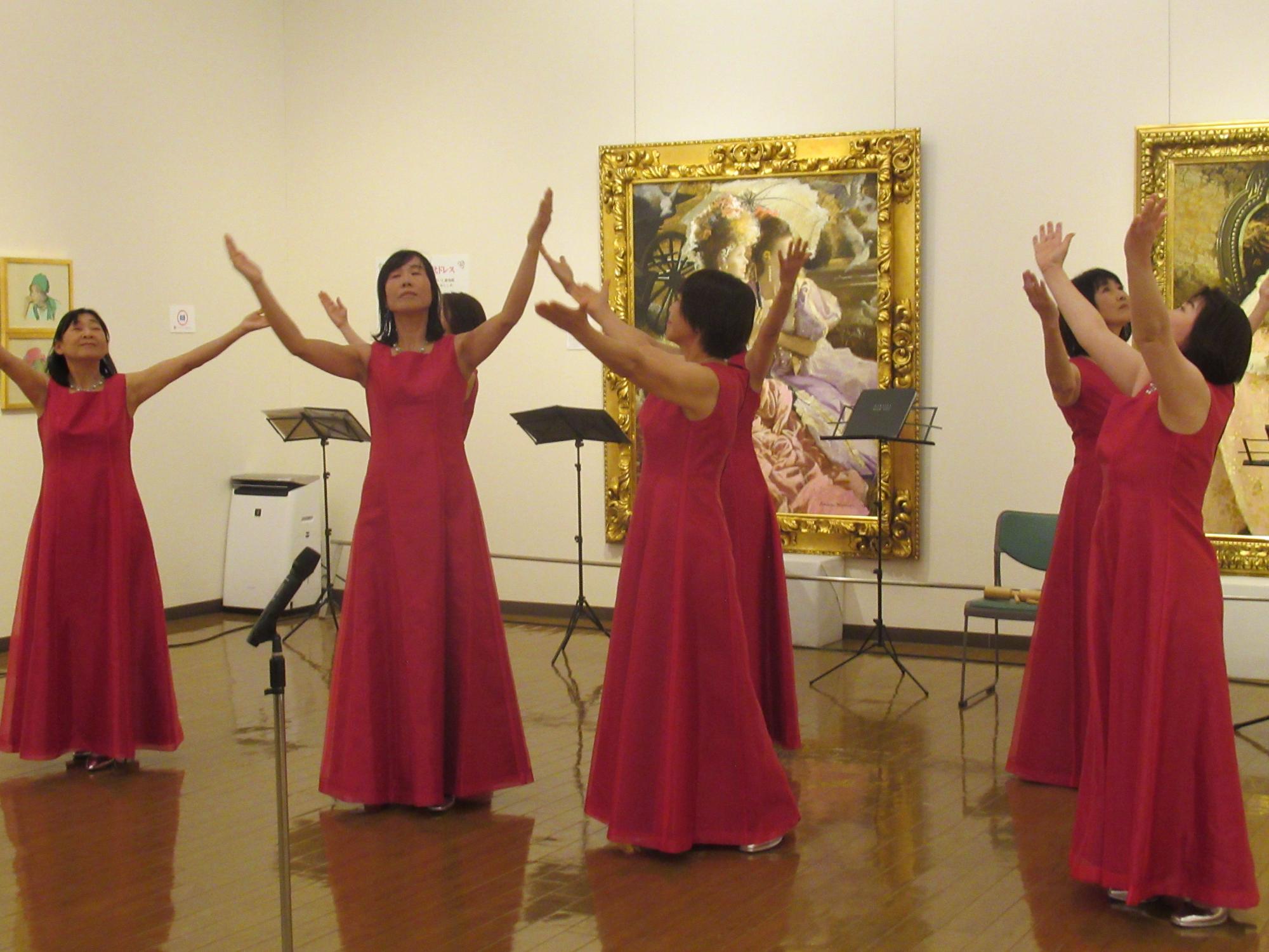 背後には豪華な金色の額縁の大きな絵画が飾られている美術館の展示室で、赤いロングドレスを着た7人の女性たちが両手を高く上げてポーズを取り歌を披露している一場面の写真