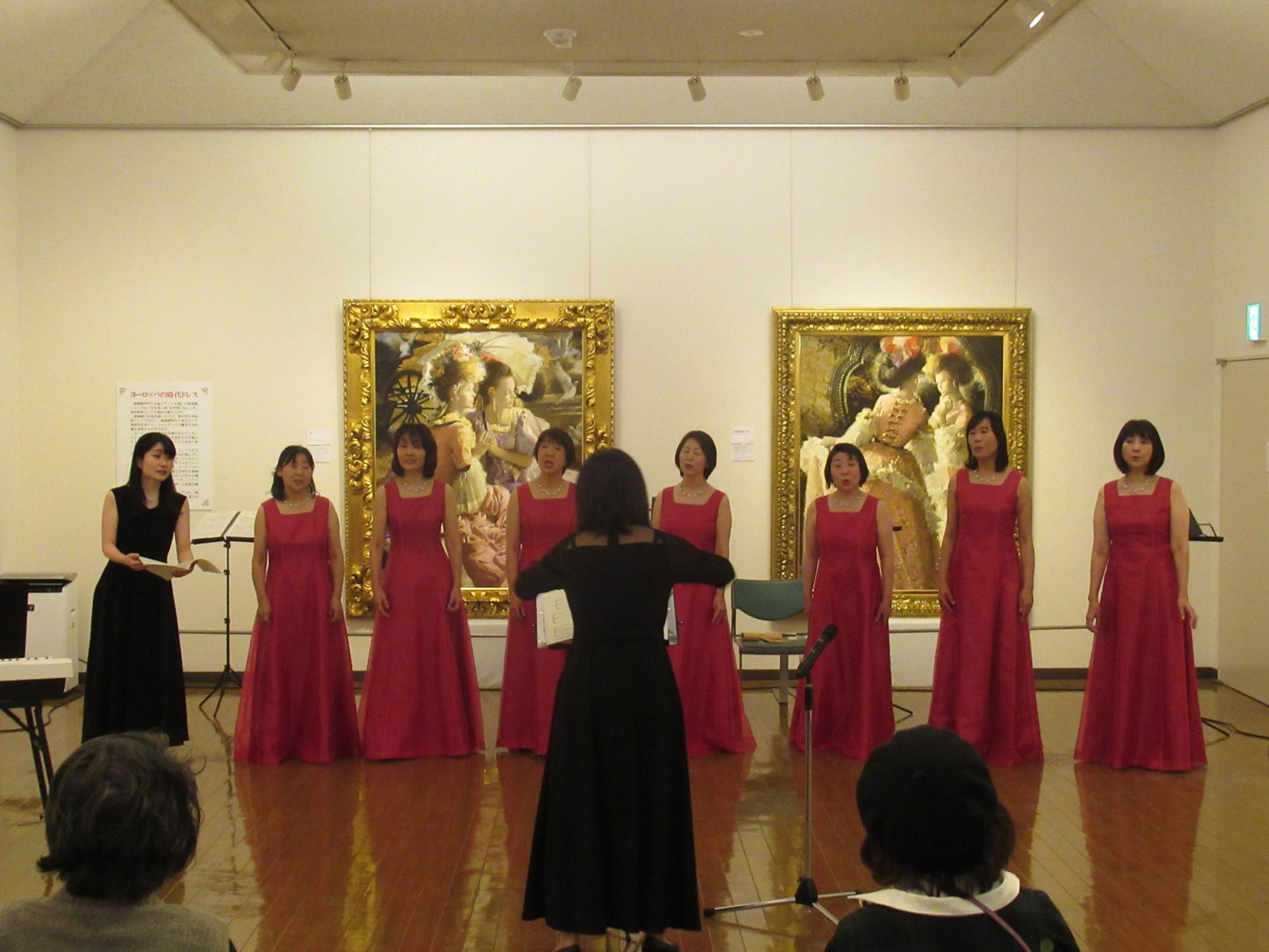 豪華な額縁に入った絵画が飾られた展示室で、中央の黒い衣装の指揮者に合わせて、向かい合って立つ左端に黒いドレスの伴奏者が立っており、その横で赤いロングドレスを着た7人の女性が横一列に並んで歌っている様子の写真