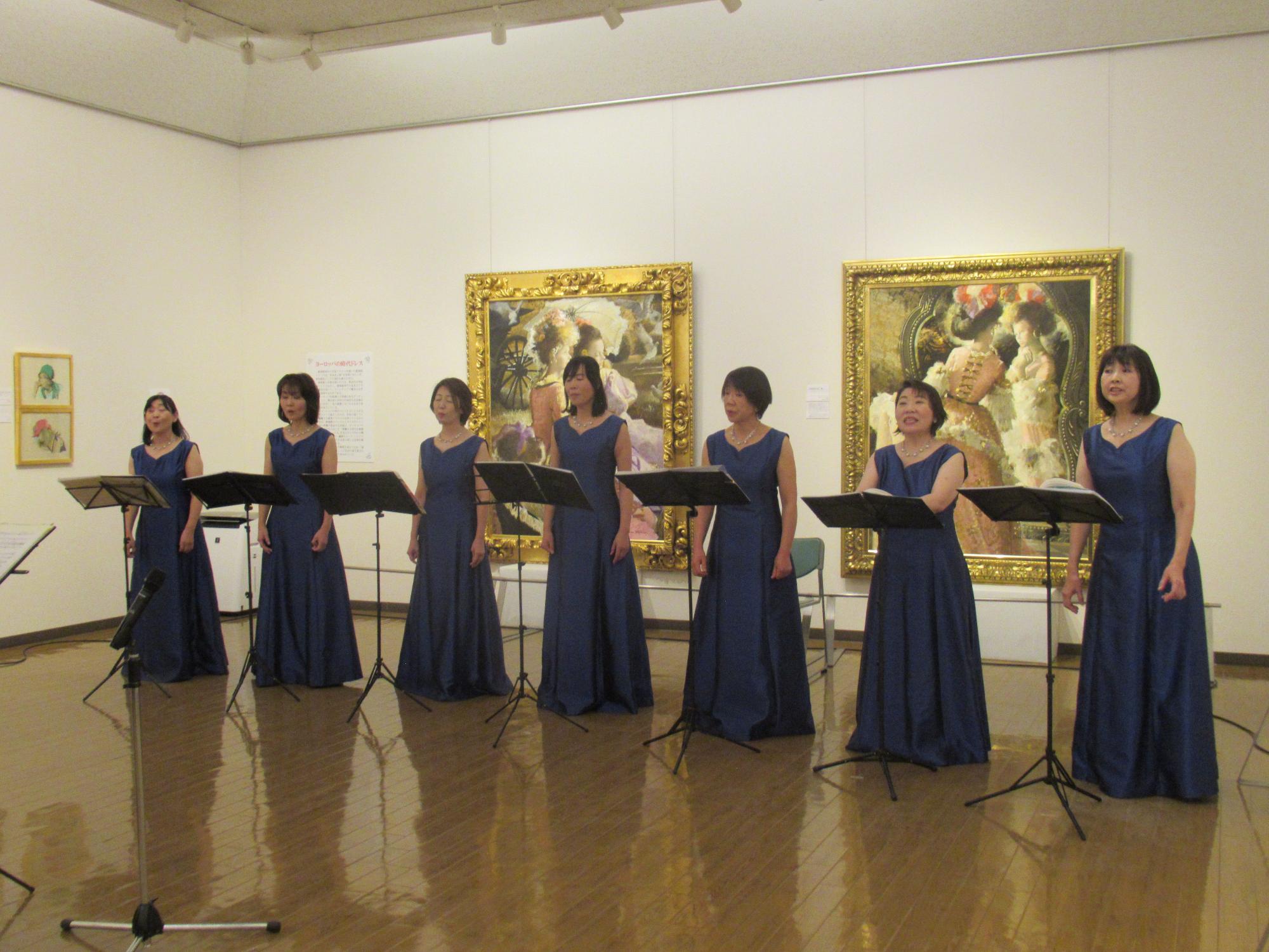 豪華な額縁に入った油絵が飾られた白い壁の展示室で、濃紺のノースリーブのロングドレスを着た7人の女性合唱団員が譜面台の前に立って歌っている様子の写真