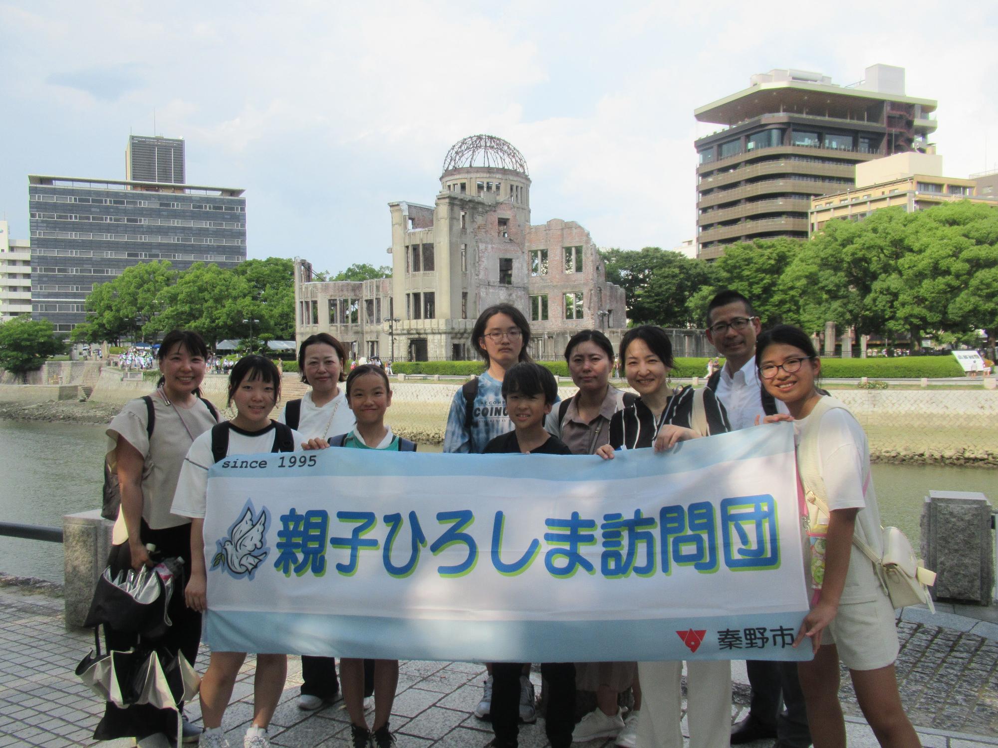 原爆ドームが見える太田川沿いの平和記念公園で、様々な年齢の男女十人が「親子ひろしま訪問団」と書かれた横断幕を手に笑顔で並んでいる記念写真
