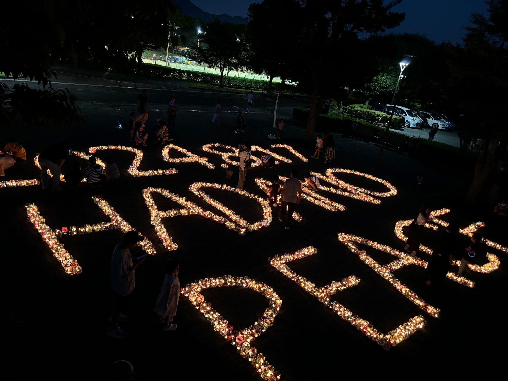 夜の公園の暗い地面に多数のキャンドルで「2024年8月17日HADANO PEACE」という文字が形作られており、周囲にはその光景を見つめる数人の人々が立っている写真