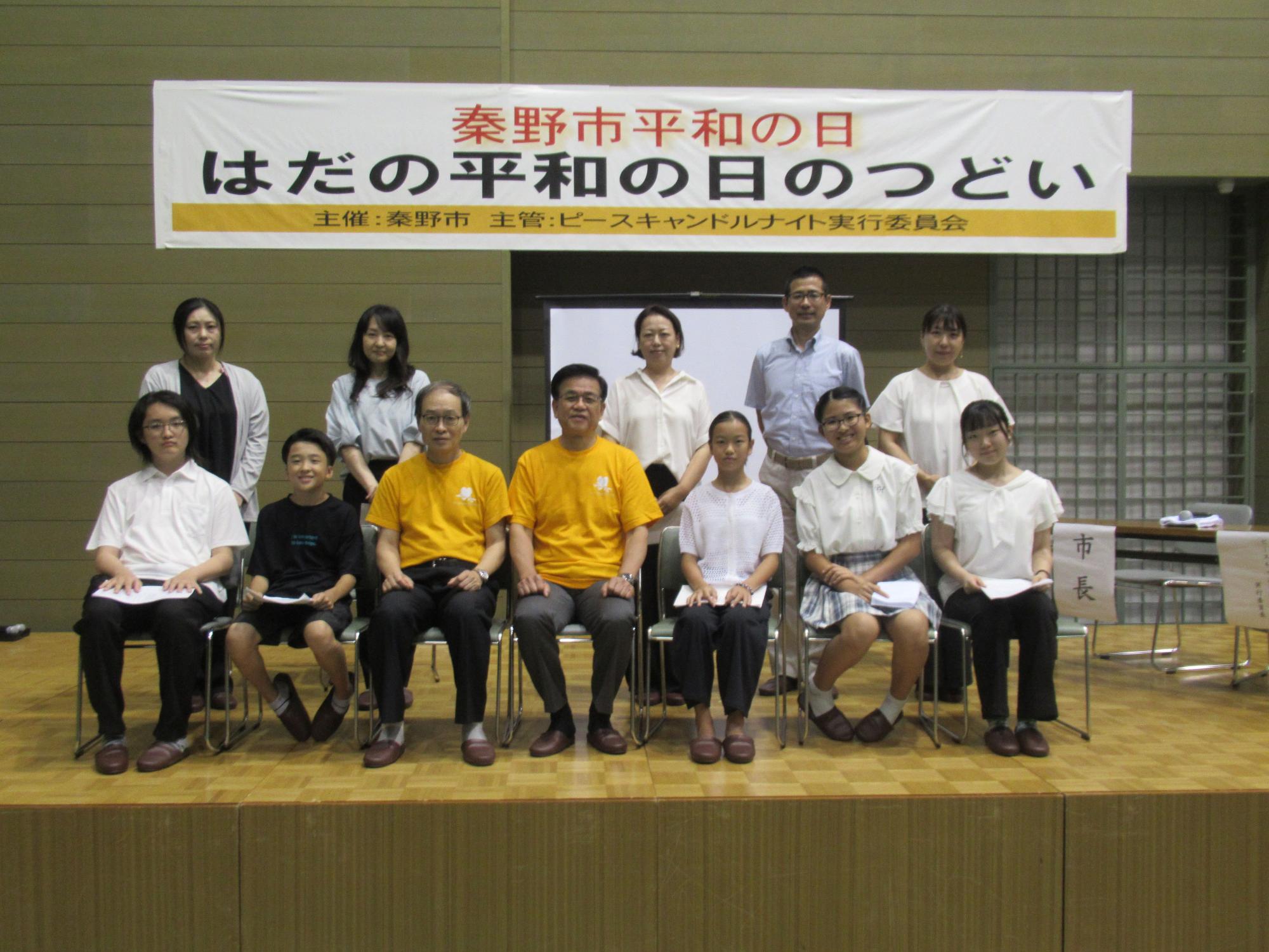 横長の「秦野平和の日 はだの平和の日のつどい」と書かれた横断幕が掲げられた講堂のような場所の舞台上で、白や黄色のシャツを着た複数の人達が立ったり椅子に座ったり2列に並んでカメラに向かって微笑んでいる集合写真