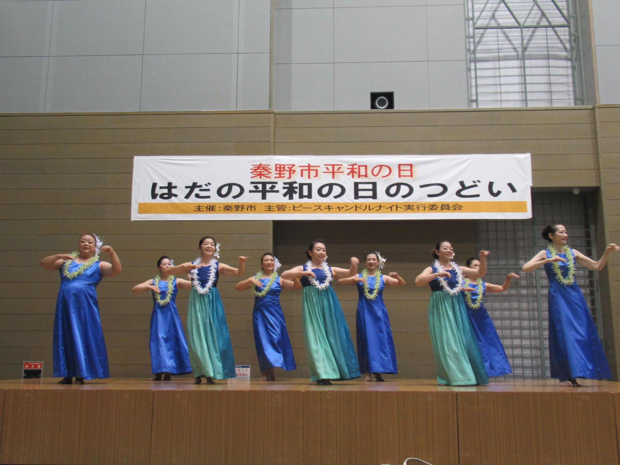 「秦野平和の日 はだの平和の日のつどい」の横断幕が掲げられたホールのような舞台上で、青と緑色のロングドレスを着てレイをかけた女性9名が、フラダンスを踊っている場面を捉えた写真