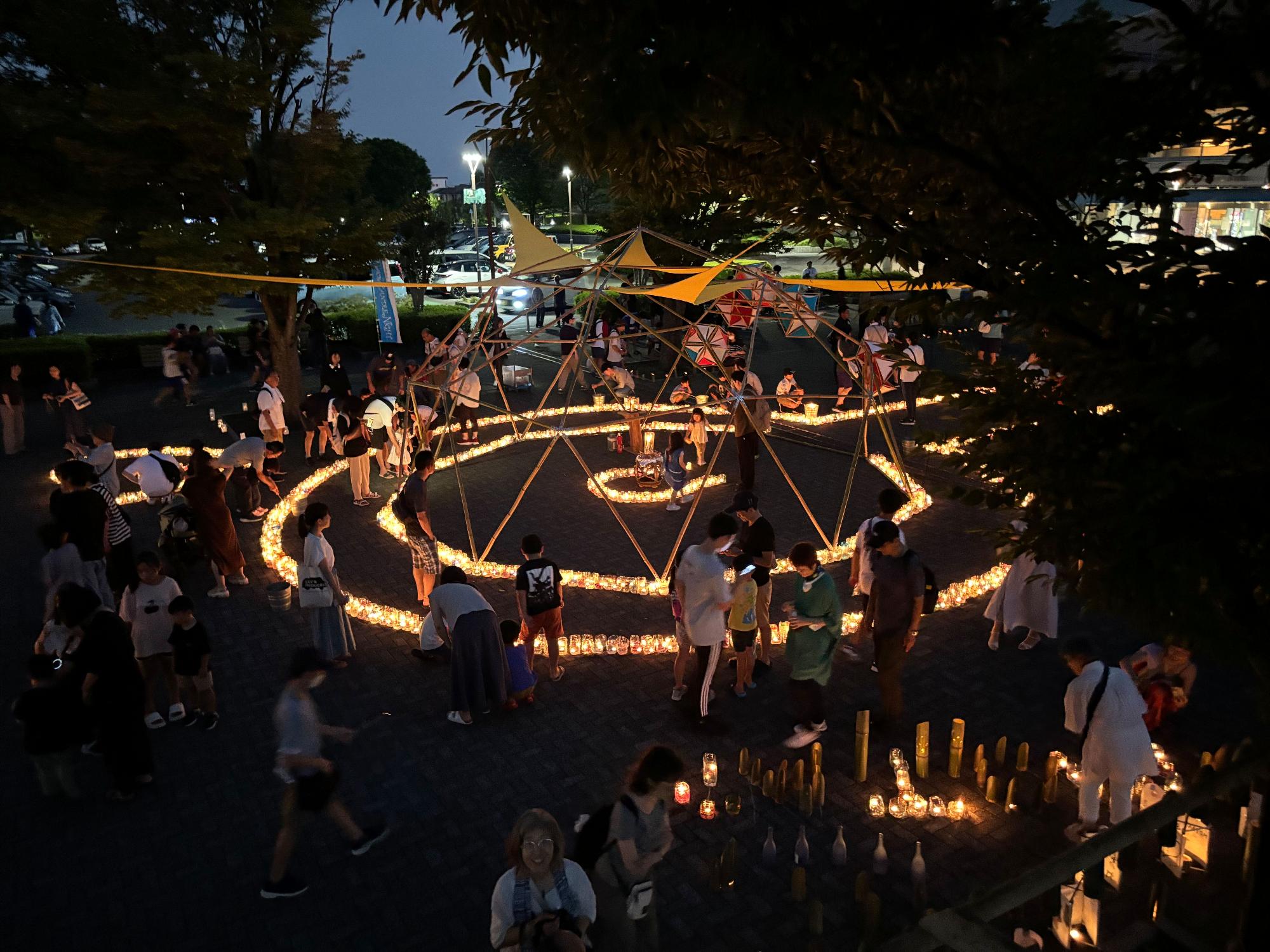 夜の屋外の広場で、無数のキャンドルが丸い幾何学的な形に配置され、その幻想的な光の中を多くの参加者が歩き回ったり見つめたりしているイベント風景の写真