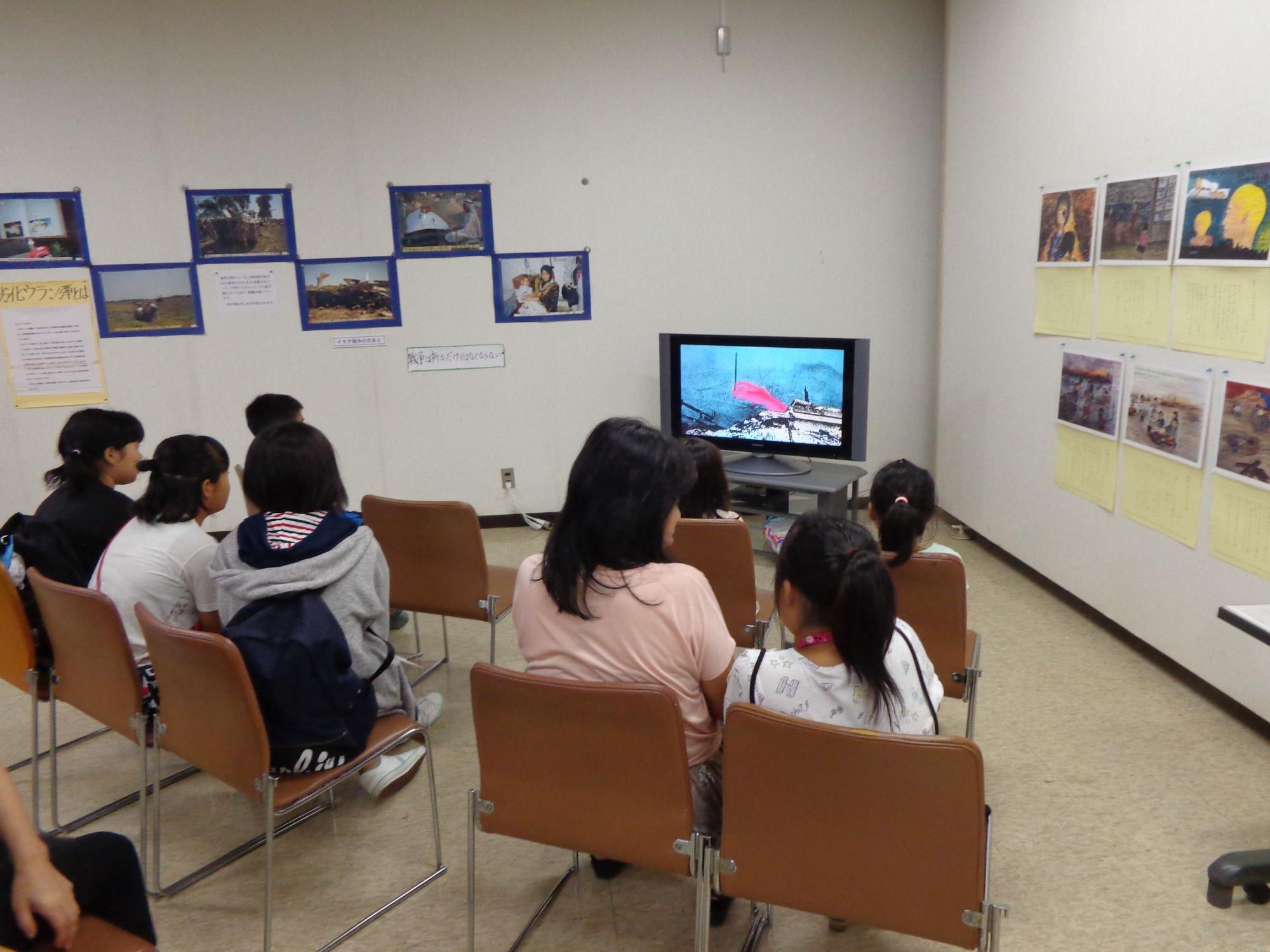 茶色の椅子に座った親子連れの参加者が、壁に展示された写真や黄色い紙の掲示物、左隅の白い掲示物がある部屋で、モニターに映し出された映像を集中して見ている後ろ姿の写真