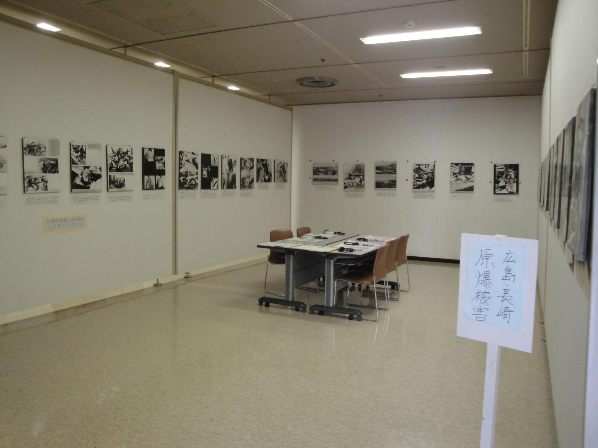 「広島・長崎 原爆被害」と手書きされた白い立て看板が手前に設置された部屋の中央には5脚の茶色の椅子が置かれたテーブルがあり、壁沿いに戦争の被害の様子を撮影した白黒の写真が展示されている展示室の写真