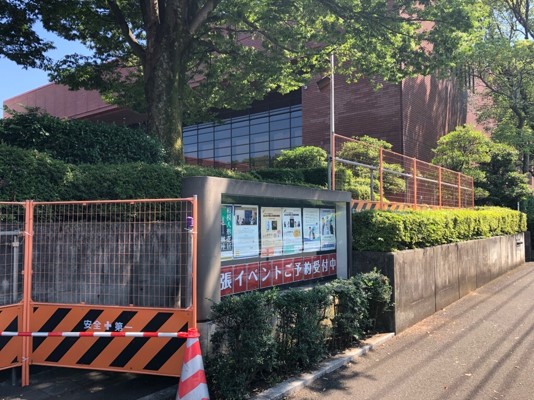 大きな木と手入れされた植え込みの奥茶色のレンガの建物が見え、手前の施設内のイベントポスターが貼られた掲示板を両側に「安全第一」と書かれたオレンジと黒の工事用バリケードで敷地を囲んでいる写真