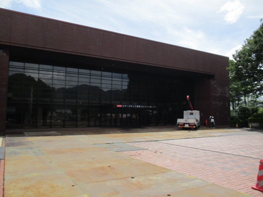 正面全体が黒いガラス窓となっている重厚な茶色のレンガ造りの外壁を持つ大きな建物の入口前の広場の赤レンガの床の上に錆びた鉄板が敷かれ、高所作業車と作業員が写っている工事の様子の写真