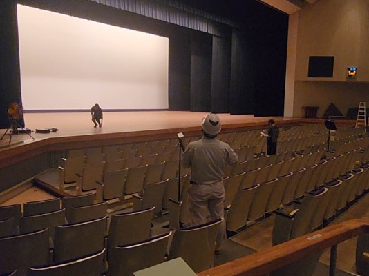 白い巨大なスクリーンが設置されたステージ上で作業する人、客席で作業員が音響測定を行っている作業中の様子をホールの客席後方から捉えた写真