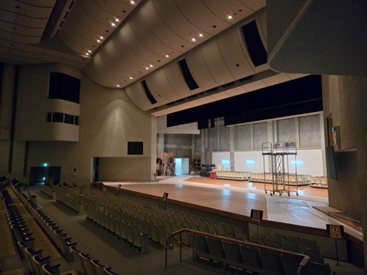 手前には空席の客席が並び、カーブした天井と反射板の下の舞台上には椅子が数脚と作業用の足場が置かれている、大きなコンサートホールの工事中の様子の写真