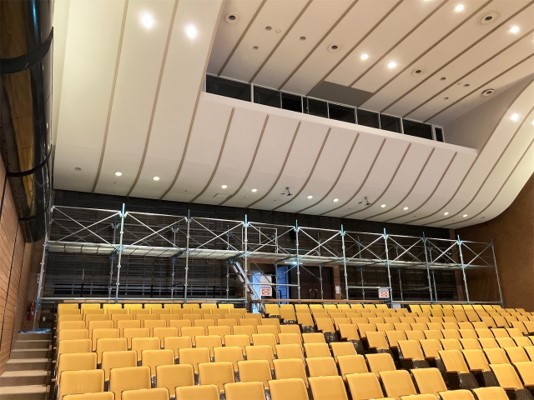 オレンジ色の空席が並ぶ上に、照明が埋め込まれた緩やかな曲線を描く白い天井と、音響や照明の操作盤が配置されていると思われる上部をの下に、大規模な金属製の足場が組まれている写真
