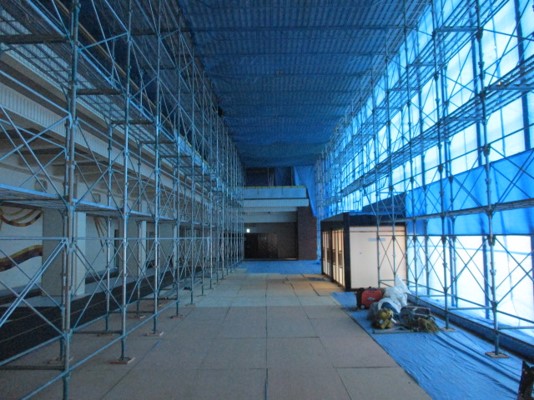 床は養生ボードで覆われ、天井と窓は青い養生シートで覆われ、通路の両側には天井まで届く大規模な金属製の足場が組まれている大ホールの全面改修中の様子の写真