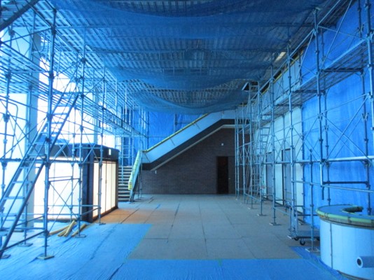 床と壁の一部が青い養生シートと養生ボードで覆われ、天井まで届く足場が組まれた空間の中央に、上階へと続く階段と、その下に壁とドアが見える、ロビーの改修工事現場を写した写真