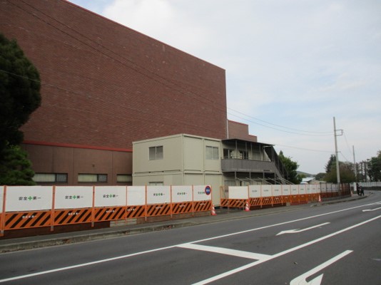 車道側の敷地内にレンガ調の大きな建物が立ち、手前に設置された「安全第一」と書かれた白いパネル付きのオレンジと黒の工事用バリケードと、その内側に置かれたクリーム色の二階建てのプレハブ事務所が写っている写真