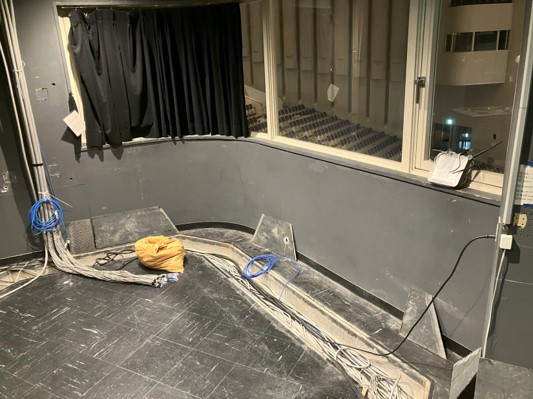 黒いカーテンが引かれた暗い部屋の一角に客席が見える窓があり、その床にはケーブルを収める溝と複数の青いケーブルや太いケーブルが無造作に置かれており、床や壁はグレー色で統一されている音響室の写真