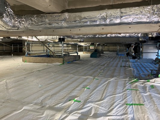 銀色の断熱材で覆われた大きなダクトが通るコンクリートの梁の下に、床一面が白い養生シートで覆われ、中央には免震装置または床構造を構築するための基礎のようなものが組まれている撤去作業中の天井裏の様子を撮影した写真