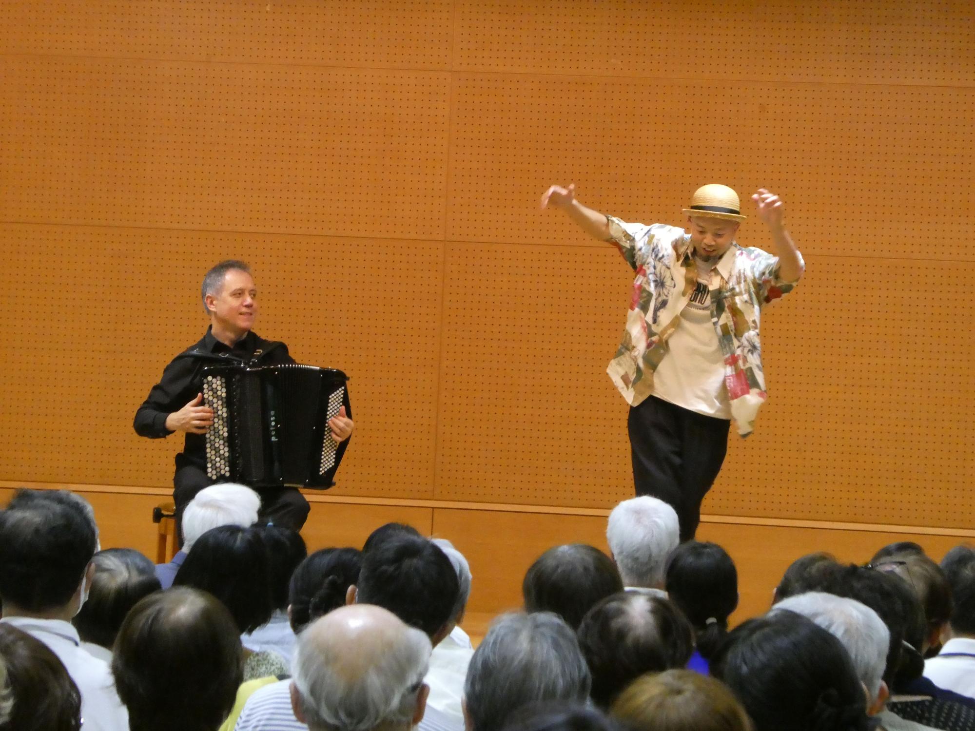 ステージ上で左側に座る黒いシャツの男性のアコーディオンの演奏に合わせて、右側で麦わら帽子とアロハシャツ風の衣装の男性パフォーマーが両手を広げて踊っている様子の写真