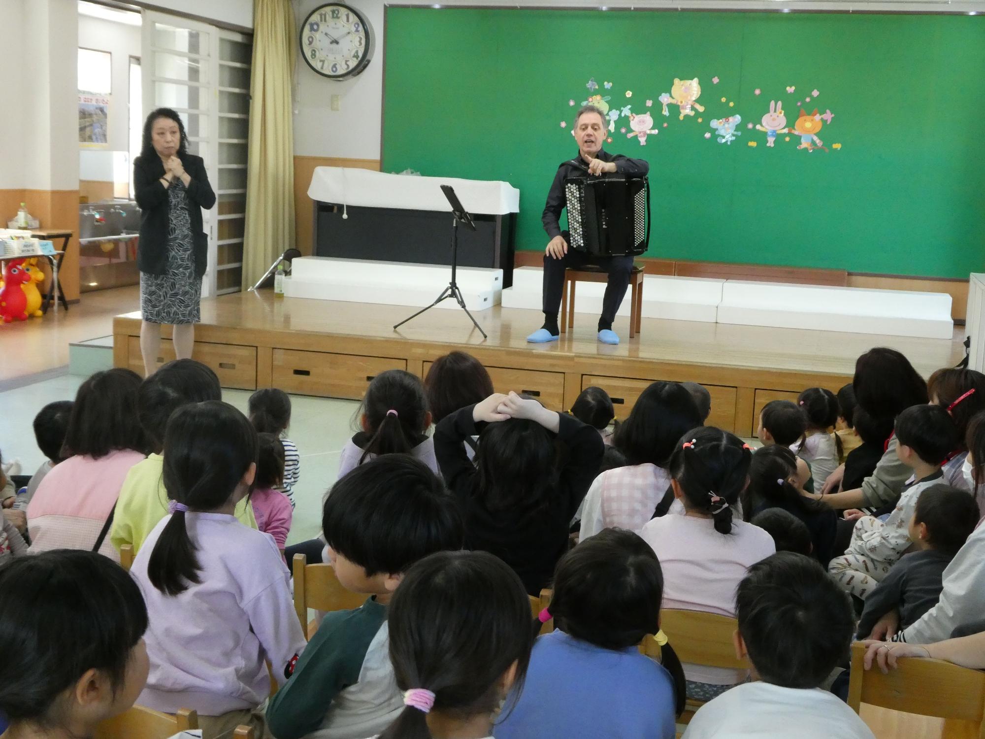 可愛らしい動物のイラストが貼られた緑の壁の背景に、黒い服の女性が立ち、中央の小さなステージの椅子に座った黒い服を着たアコーディオン奏者の男性が向かい合って座っている園児たちに向けて話しかけている写真