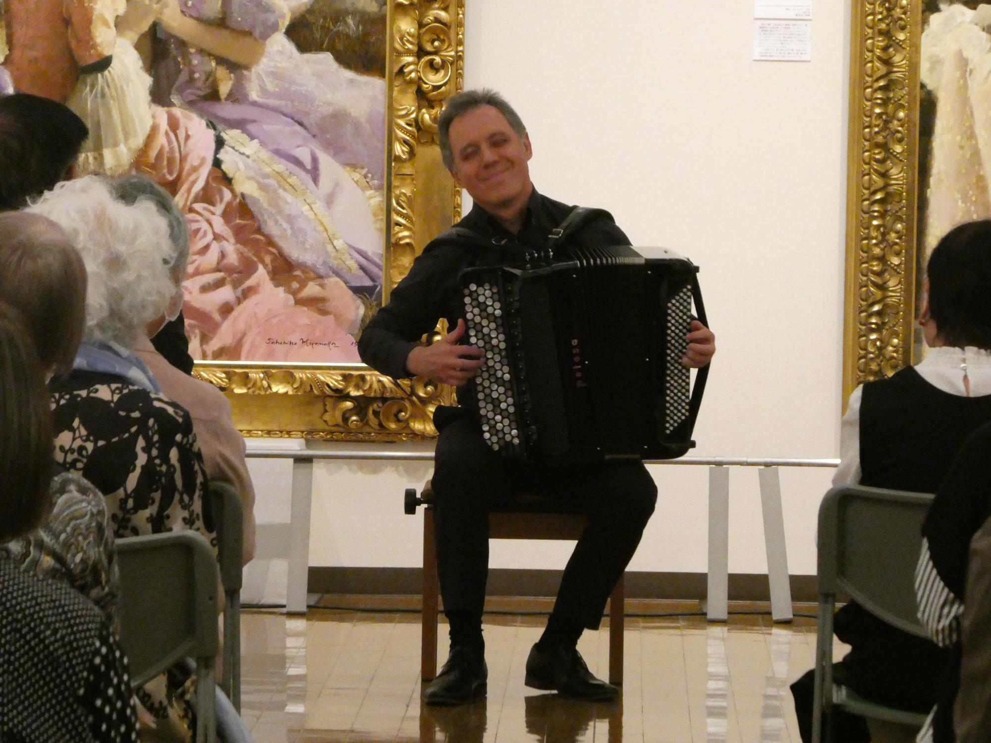 金色の額縁に入った絵画が飾られた美術館の展示室で、黒い服を着たアコーディオン奏者の男性が椅子に座り、聴衆に笑顔を向けながら演奏している様子の写真