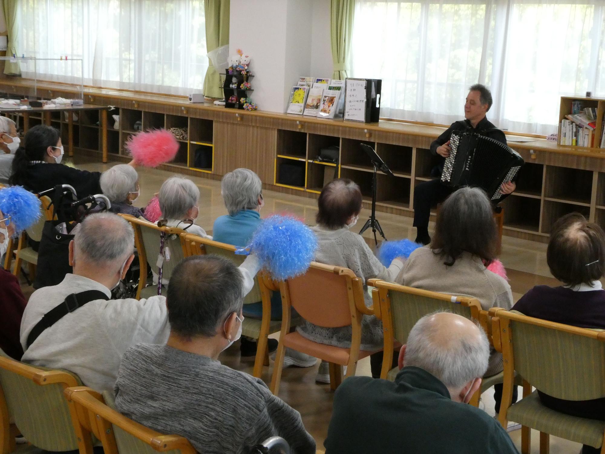 施設の一室で、一部の観客は青やピンクのポンポンを持っている高齢の観客が椅子に座り、中央前方に座ってアコーディオンを演奏する黒い服の男性奏者に熱心に聞き入っている様子の写真