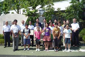 建物の前に立つ木々に囲まれたモニュメントの前で、大勢の子供と大人が3列に並んでおり、全員がカメラの方を向いて立っている集合写真