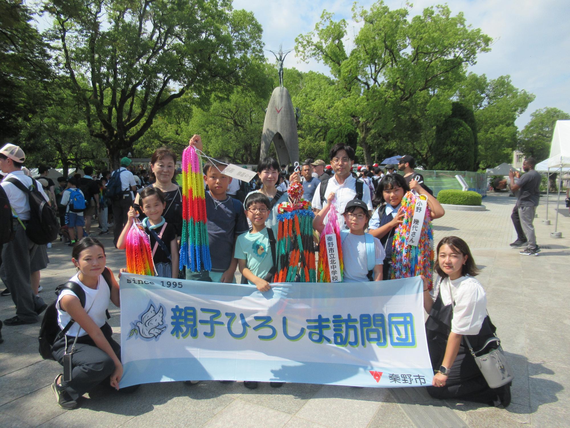 平和記念公園で、様々な年齢の男女十人が「親子ひろしま訪問団」と書かれた横断幕と千羽鶴を手に笑顔で並んでいる記念写真