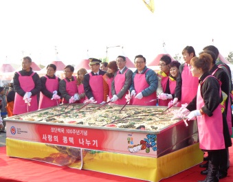 ピンク色のエプロンを着用した関係者達が巨大な四角い容器の中を長い柄のへらで混ぜているセレモニーの様子の写真