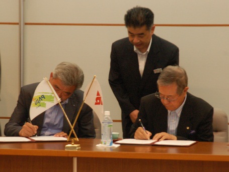 中央に日韓友好の意匠が施された旗が立てられたテーブルで、チャン・ドンムン会頭と関野義一会頭が書類に署名している様子を、後ろに立つ男性が静かに見守っている調印式の写真